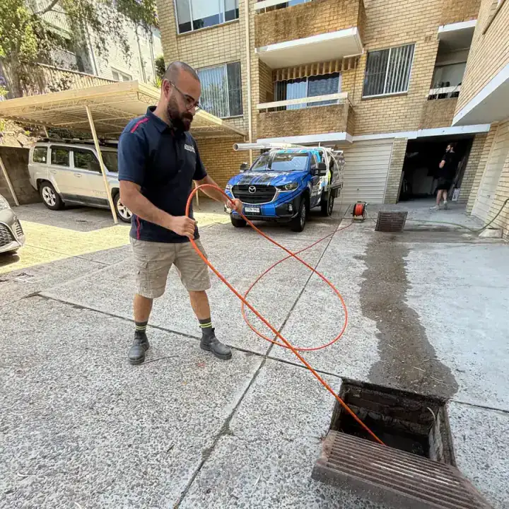 Antons plumber feeding a drain hose into an open pit beside an apartment building
