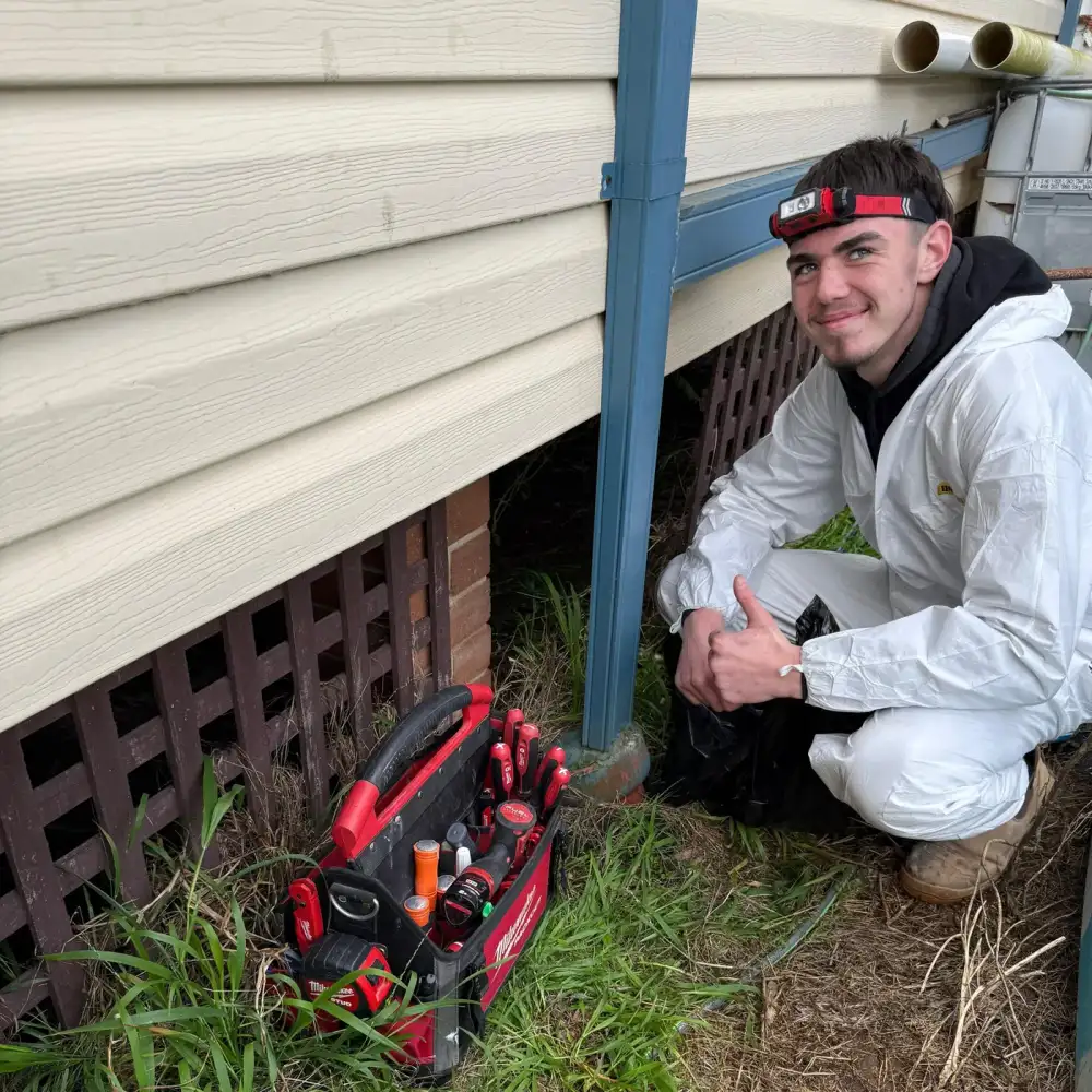Technician with a headlamp and tool bag beside a subfloor access opening